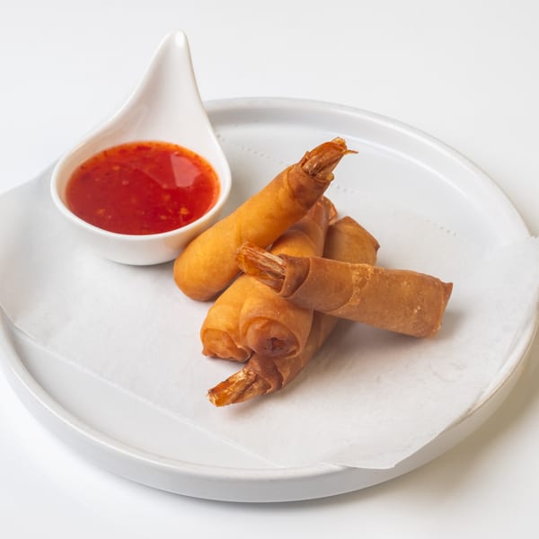 Crispy KorKai Thai shrimp spring rolls with a side of sweet chili dipping sauce on a white plate.